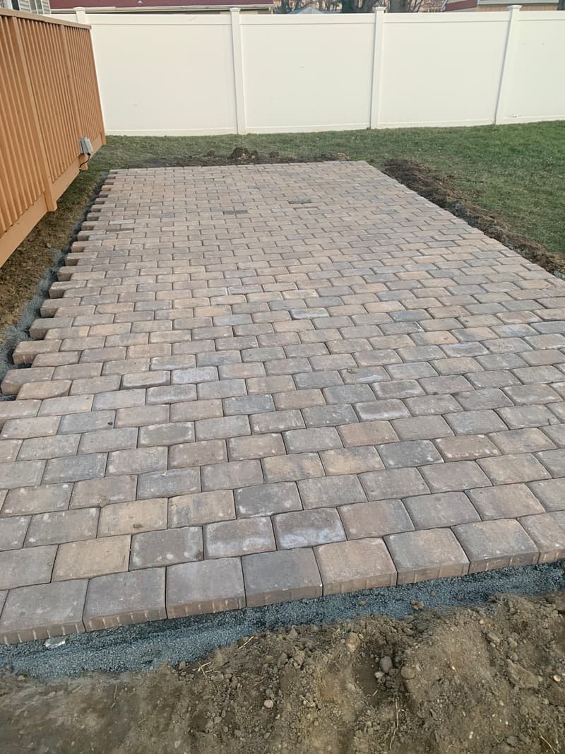 Newly laid tan and brown paver patio with a border course, next to a deck stair and a white vinyl fence
