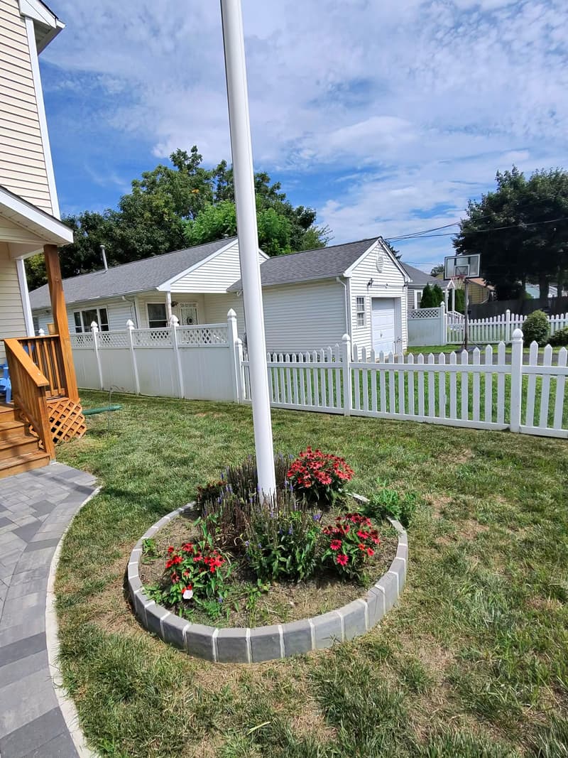 Gray paver walkway curving past a flagpole bed ringed in paver blocks and planted with red flowers and purple salvia
